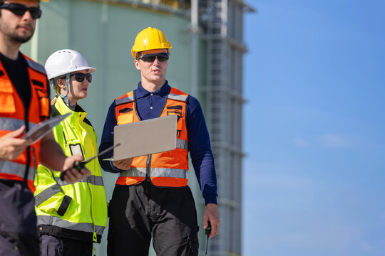 Industrial engineers with laptop and walkie talkie inspecting gas tank facility outdoors, Engineering team wearing hard hats and safety vests discussing project at refinery plant