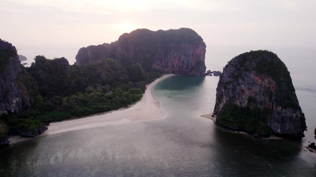 Backlit aerial drone shot of a narrow sandy beach nestled between two dark limestone cliffs with silvery water and a hazy sky in southern Thailand