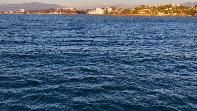 A wide aerial shot capturing a whale surfacing in the deep blue waters of Manzanillo, Colima, with a coastal town and mountains in the background during golden hour.