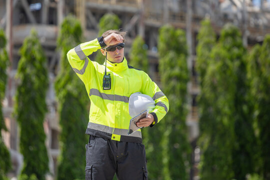 Exhausted industrial engineer wiping sweat from forehead outdoors at construction plant, Tired factory worker wiping forehead during hot weather inspection at industrial facility
