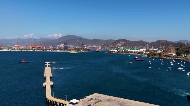 Wide aerial shot of the busy industrial shipping port in Manzanillo, Colima, featuring tugboats, cargo terminals, and mountain landscapes under a clear blue sky.