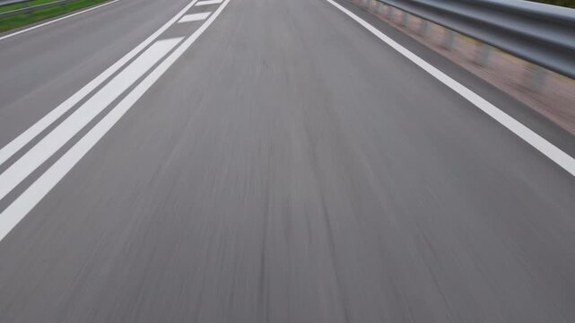 The road surface and the white lane lines blur under the car with the crash barrier along the edge. Filmed from a moving car, low angle on the road.
