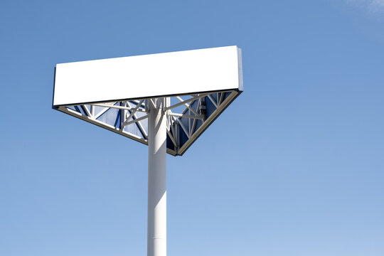 A blank white billboard mounted on a tall steel pole with a triangulated support structure against a clear blue sky. A clean empty advertising space ready for commercial advertisement mockup or design