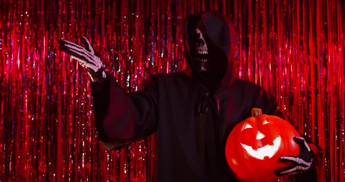 Male person wearing Halloween death costume and skull mask having fun blowing a kiss on festive red foil background holding pumpkin celebrating holiday. Halloween party and horror concept. 4k video.