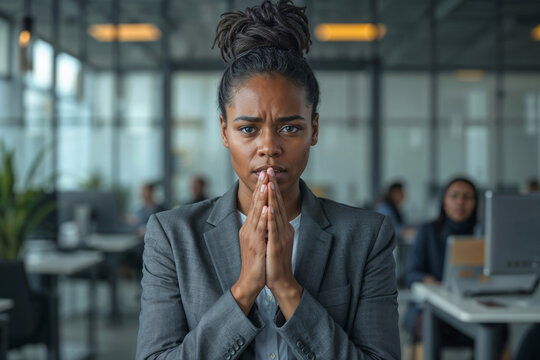 Stressed Black woman in office, hands clasped in prayer. Upset office worker black woman, clasping her palms together, pleading not to be fired against the backdrop of a modern office