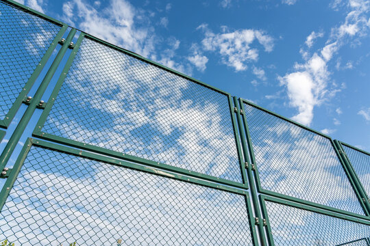 A green metal wire mesh fence viewed from a low angle, set against a bright blue sky with scattered white clouds.