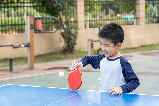 A young asian boy plays table tennis happily at an outdoor sports ground, with the ping pong ball in mid-air.