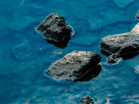 Dark mossy rocks emerging from clear deep blue water with submerged stones visible below the surface, calm river or pond nature scene