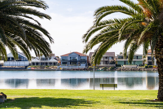 A peaceful lakeside foreshore reserve at West Lakes in Adelaide, South Australia, with modern waterfront residential homes reflected in the still lake surface. Relaxed and affluent lifestyle.