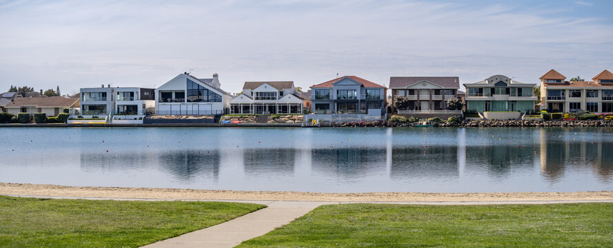 Panoramic view across the calm reflective waters of West Lakes in Adelaide, South Australia, showing a row of modern upscale waterfront residential homes. Upmarket canal estate lifestyle