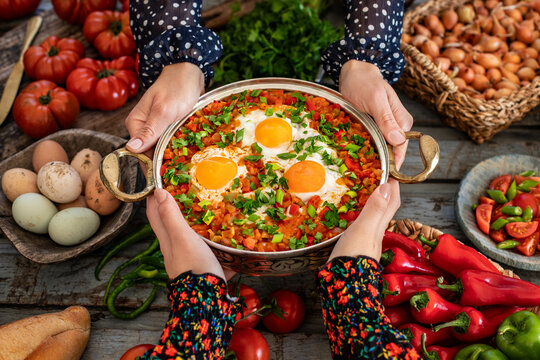 Shakshuka, fried eggs with tomatoes, onion, red pepper and spices in cast iron pan, other name is menemen. Hands of two women sharing and holding their meal.  