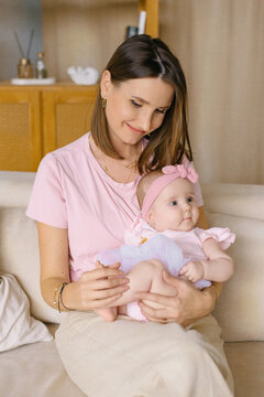 Young Mother Holding Her Cute Baby Daughter In Pink Outfit While Sitting On Sofa
