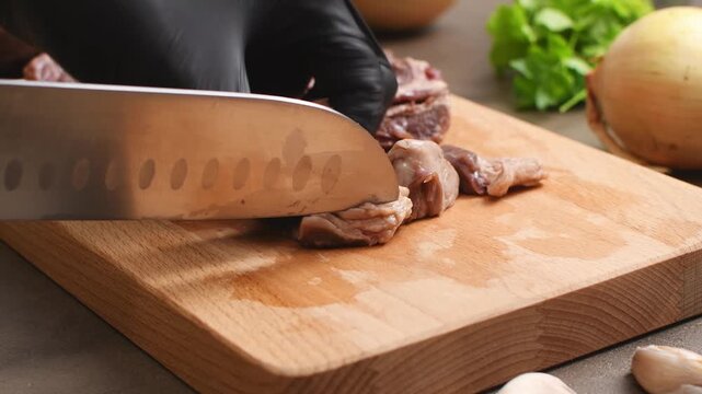 A gloved hand cuts away thick white fat from pieces of beef with a knife on a wooden cutting board.