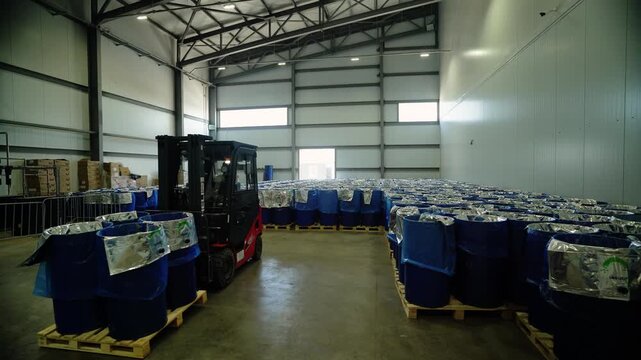 Forklift moving stacked blue barrels in large warehouse