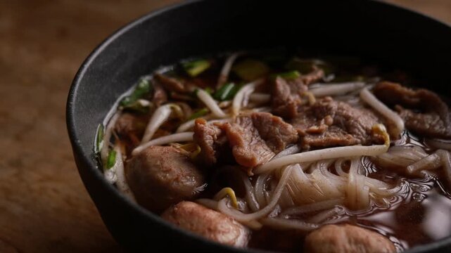 Close-up of Thai boat noodle soup with sliced beef, meatball, rice noodles, bean sprouts and green onion in rich dark broth, served in a black bowl on rustic wooden table
