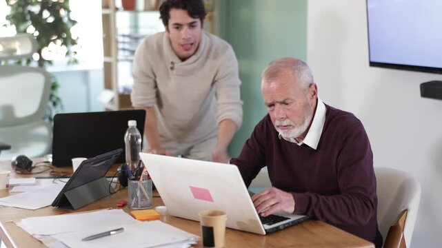 Senior man leaned over to seated guy employee, supervise activities, helps with work and looks at laptop screen. Travel agency, engineering company, notary office or legal advice 