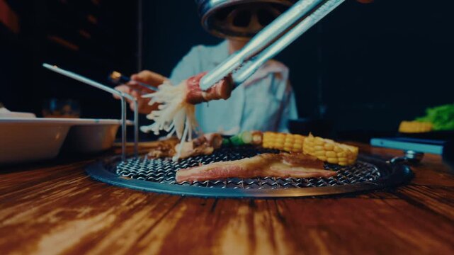 Two pairs of tongs turning bacon wrapped mushrooms and corn on a barbecue grill at a restaurant table, Da Lat, Vietnam