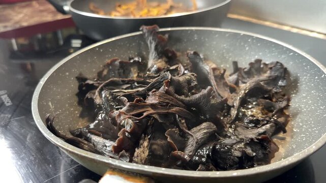 Dark Craterellus cornucopioides black trumpet mushrooms frying in a steel pan. Horn of plenty, wild mushroom cooking, foraged food, autumn harvest, seasonal recipes, gourmet ingredient.