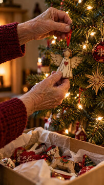 Wrinkled hands hanging a worn wooden angel ornament by a red ribbon on a Christmas tree above a tissue-lined storage box, with warm lights showing gentle care for familiar holiday decorations