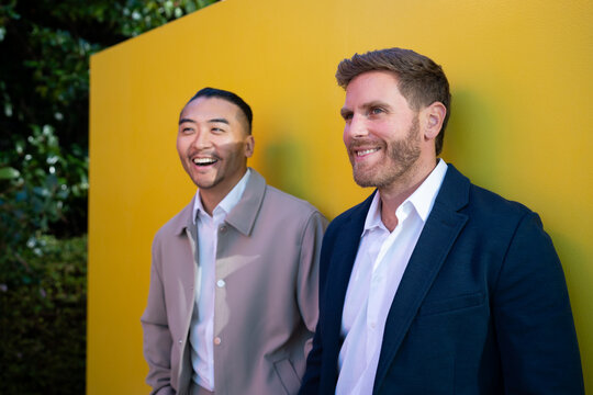 Two white men smiling outdoors against a yellow wall