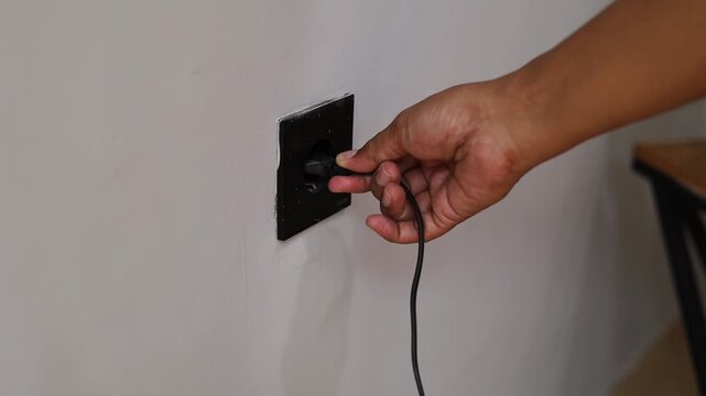 A close up view of a person's hand holding an electric plug and preparing to plug it into a wall outlet, depicting electricity, energy, home appliance usage, and safety.