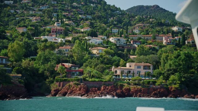 Pink villa with garden and parasols above red rocks and turquoise sea, with hillside houses and a green peak behind, Esterel, France.