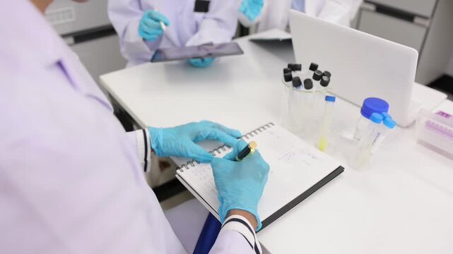 Scientific researcher in a lab coat and gloves analyzing a liquid sample in a test tube and taking notes in a notebook for chemical research.