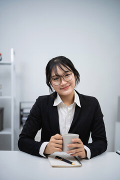Young woman in business attire holding cup sitting at white desk with notebook and pen in modern office