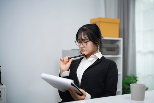 Thoughtful young woman holding pen to mouth while reading document in bright office with modern decor