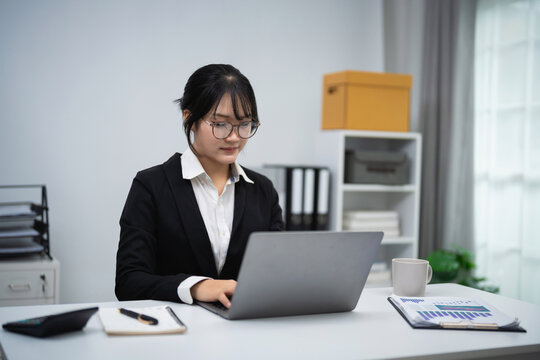 Young professional woman in black suit working focused on laptop at modern office desk with documents and coffee cup