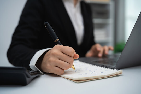 Businessperson in formal suit writing notes with pen in notebook while working on laptop at office desk