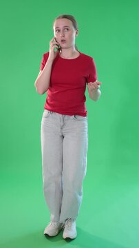 Young teen girl dressed in red T-shirt and light blue jeans talking by smartphone standing against green screen 