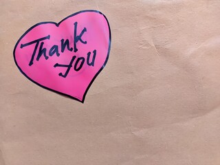 Pink heart-shaped sticker with handwritten text "Thank You" on a brown paper background © Putu