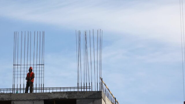 A builder wearing orange safety gear surveys a rooftop lined with steel rebar