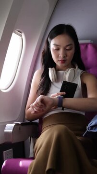 Asian tourist woman using a smartphone on an airplane flight. Young Asian woman checking her smartwatch and using her smartphone while traveling by airplane, sitting next to the window seat.