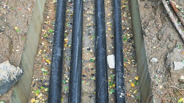 Black pipes lie lengthwise in a gravel trench beside wooden edging, with small plants and soil around them. Their arrangement remains steady as the camera view shifts slightly over time