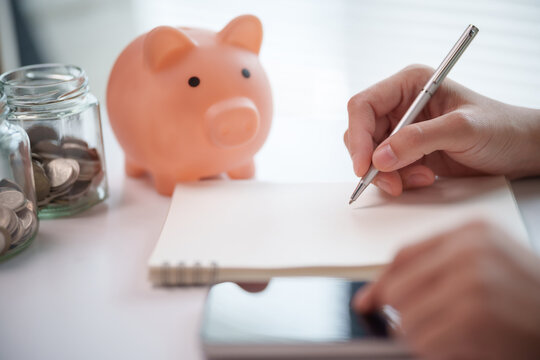 Close-up of a hand writing in a blank notebook beside a pink piggy bank and glass jars filled with coins on a desk, represents savings planning, financial goals and household budgeting.