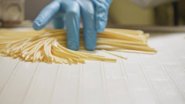 A blue-gloved worker gathers long pasta strands into a bundle on a white conveyor at a food production workstation.