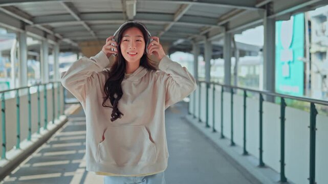 Happy young Asian woman putting on wireless headphones and dancing joyfully to music on modern urban pedestrian walkway, smiling female enjoying rhythm with arms raised, freedom,