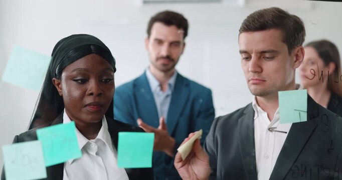 Portrait of marketing professionals offer and discuss insights in board office. Multiethnic managers put memo stickers onto glass wall at group brainstorm