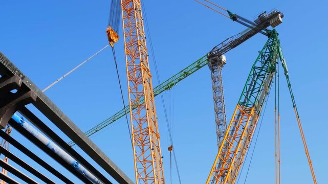 Low angle view of heavy-duty tower and crawler construction cranes operating against a clear blue sky. Industrial mechanism of multi-colored steel lattice booms and lifting equipment on a building sit