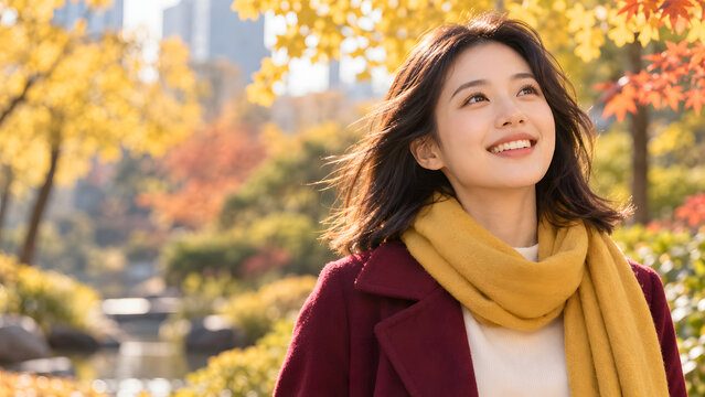 Smiling woman wears a mustard yellow scarf and burgundy coat outdoors beneath bright autumn leaves, looking upward as sunlight filters through colorful trees along a leafy path in warm light.