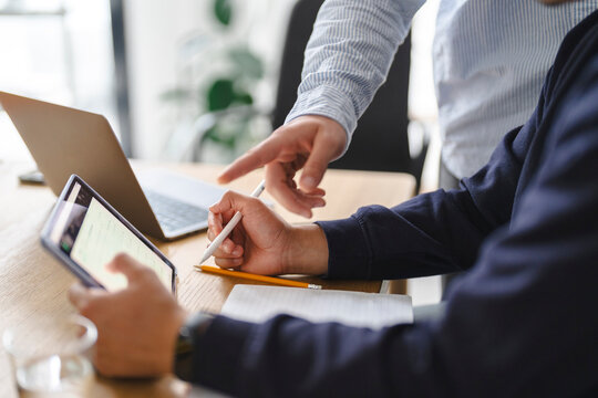 Two colleagues collaborate in a modern office. One person points at a tablet while the other takes notes with a pen, reviewing project data together at a wooden desk.