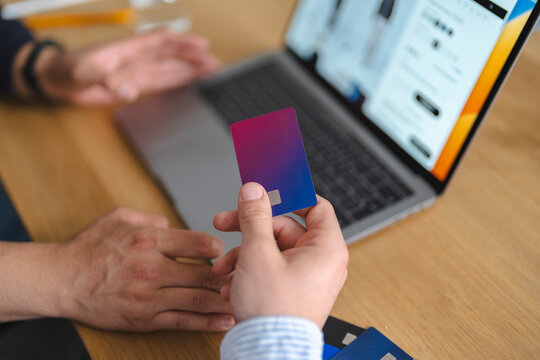 A man holding a credit card in his hand while shopping online with a laptop computer at a wooden table. He is making an online payment for his purchases.