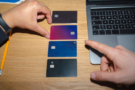 A person sitting at a wooden desk compares several plastic payment cards next to a laptop computer. One hand points at the cards while the other touches one.