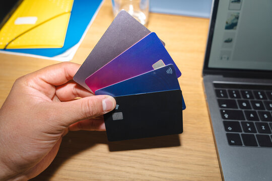 A person holding a fan of multiple plastic payment and debit cards in their hand. A laptop rests on the wooden desk next to them while they handle online shopping.
