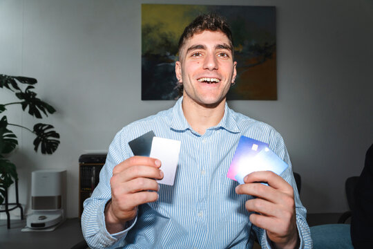 A smiling young man wearing a blue striped shirt holds multiple bank cards in both hands. He is indoors, happily showing off his new credit cards to the camera.
