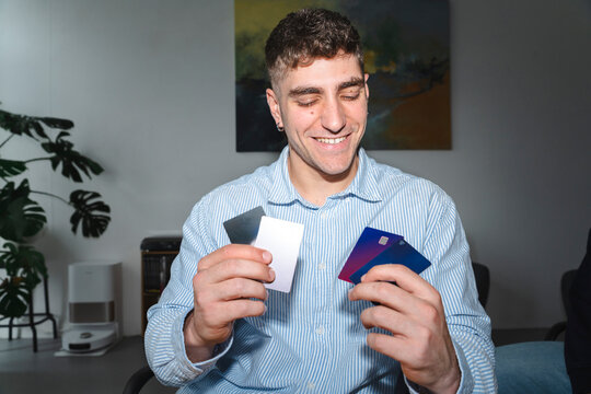 A young smiling man in a striped shirt holds several credit cards in his hands indoors. He looks down at the plastic payment cards with a happy expression in a modern room.