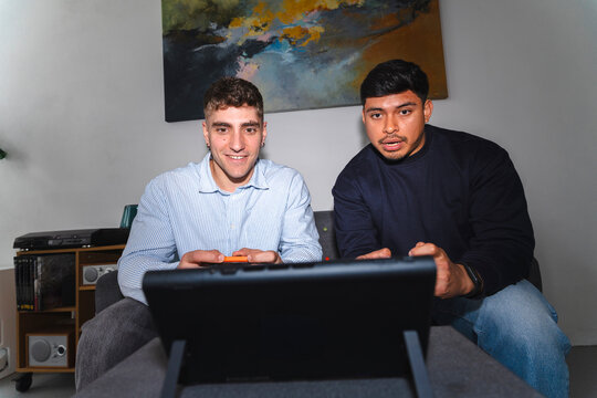 Two young men sitting on a couch in a living room, playing a video game together on a portable console. They are smiling and focused on the screen, enjoying their free time.