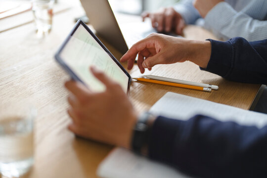 A businessman holds and reviews information on a digital tablet during a collaborative meeting in a modern office. Coworkers work together at a wooden table with laptops.